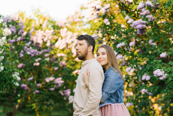 Cute Girl uściski facet z tyłu, patrzą na odległość w parku z liliowy wiosną. stosunki miłosne mężczyźni i kobiety - Zdjęcie, obraz