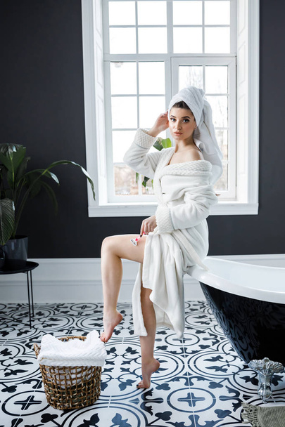 Jeune jolie fille en robe de chambre blanche et une serviette sur sa tête assise sur le bain et se rasant les jambes
 - Photo, image