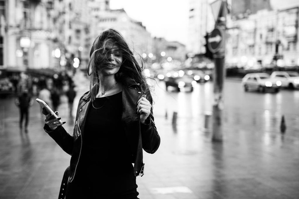 Joven chica bonita caminando por la calle con un vestido y una chaqueta, con un teléfono en las manos
 - Foto, Imagen