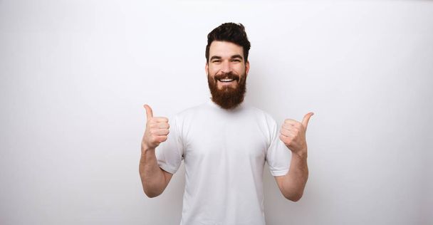 Portrait d'un homme barbu beau et excité montrant les pouces au-dessus du mur blanc
 - Photo, image