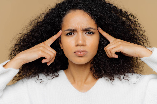 Headshot de mujer afroamericana estresante mantiene los dedos índice en las sienes, sufre de dolor de cabeza frunce el ceño, tiene expresión facial insatisfecha, vestida casualmente, modelos de interior. Personas y migraña
 - Foto, imagen