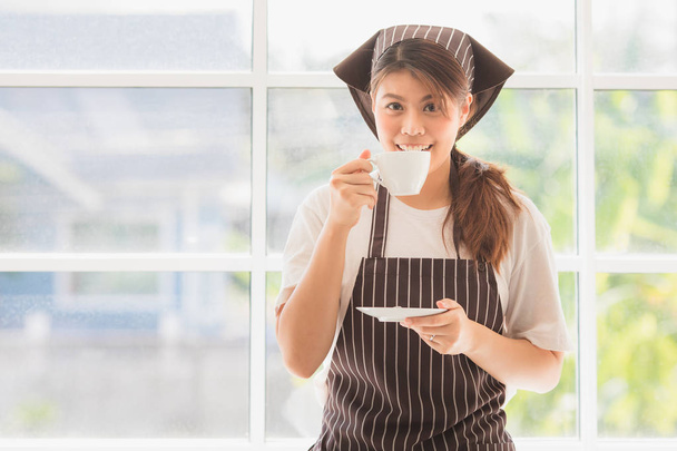 Fronte ritratto di felice giovane e bella signora asiatica a strisce cappello da cucina e grembiule, godere di pausa caffè in fondo cucina moderna con grande finestra di vetro, sorridente alla macchina fotografica
 - Foto, immagini