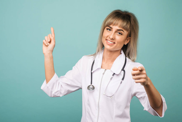 Усміхнена молода жінка-лікар зі стетоскопом вказує вверх і в камеру пальцями
 - Фото, зображення