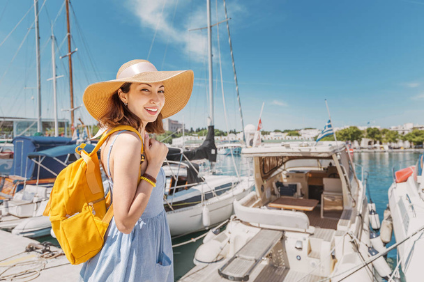 豪華なヨットと海の港の近くに立ってバックパックと帽子の若い幸せな女性。旅行と休暇のコンセプト - 写真・画像