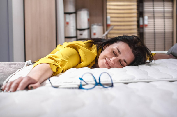 A beautiful girl is testing a new mattress before buying. Portrait of a satisfied brunette lying on a new bed. The concept of Healthy sleep - Fotoğraf, Görsel