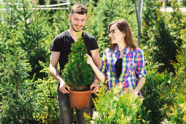 Mladý pár stojí pohromadě, drží hrnec s malým jedným stromem a dívá se na rostlinu v zahradním centru - Fotografie, Obrázek