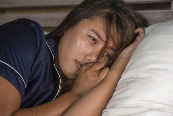 dramatic lifestyle portrait of young attractive sad and depressed Asian woman in pajamas lying in bed in pain suffering depression and anxiety feeling lonely and desperate - Foto, Bild