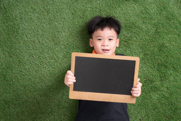 Yalan ve yazı tahtası yeşil çimenlerin üzerinde tutan şirin Asyalı çocuk - Fotoğraf, Görsel