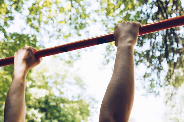 the hands of a sports athlete hold on to a metal crossbar for pull-ups on the training ground against a clear sky and green foliage. sports training outdoors. workout first-person view - 写真・画像