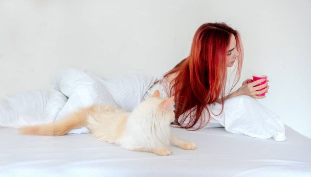 Retrato de una atractiva, contenta, joven, sexy mujer pelirroja tumbada relajada en la cama disfruta de su café y su gato yace junto a ella
. - Foto, imagen