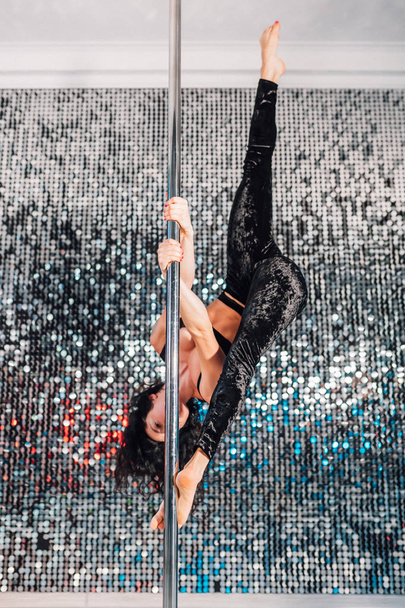 Woman doing tricks, practicing in studio with pylon. Young stripper in black dances sexually with pole in club. Poledance on shining wall background - Foto, Bild