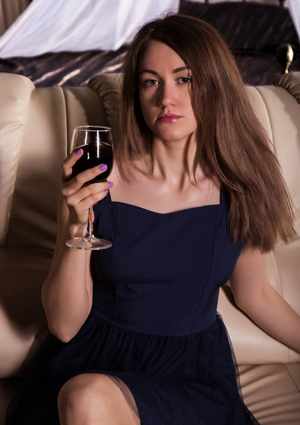 charming young woman drinks wine in hotel room, sitting on a sofa with glass of red wine - Photo, Image