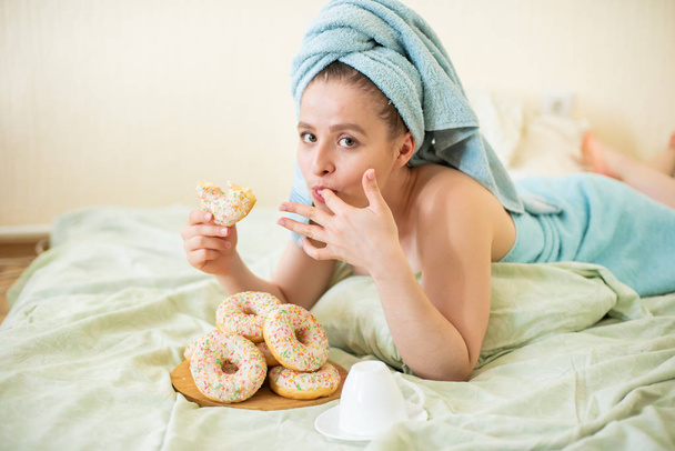 The girl in a towel has breakfast in the bed. A young attractive woman eats donuts in bed. The girl with a towel on her head happily eats donuts. - Фото, изображение