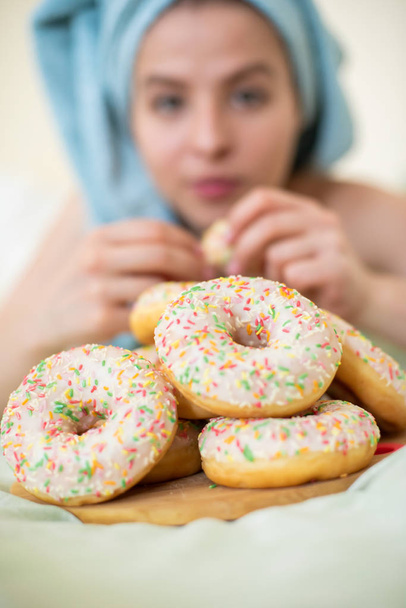 The girl in a towel has breakfast in the bed. A young attractive woman eats donuts in bed. The girl with a towel on her head happily eats donuts. - Photo, Image