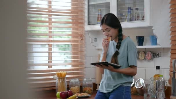  Young asian woman using tablet and eating bread. technology, social network, communication concept in kitchen at home. - Video, Çekim