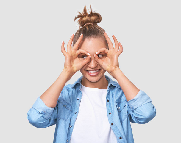Horizontal image of happy young woman with blonde bun hairstyle, showing Ok gestures with both hands, pretending to wear spectacles ot binocular. Pretty female smiling broadly and posing on white wall - Photo, Image