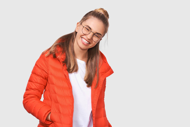 Retrato de estudio de una joven rubia feliz sonriendo boca abierta, vistiéndose de camiseta blanca y chaqueta roja, usando gafas redondas, aislada sobre fondo blanco. Concepto de personas y emociones
 - Foto, Imagen