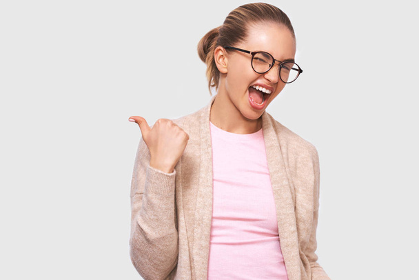 Cheerful young woman smiling broadly, dressed in casual outfit and round transparent eyewear, blink with an eye, point to copy space, likes somebody's idea, poses over white studio wall. - Photo, Image