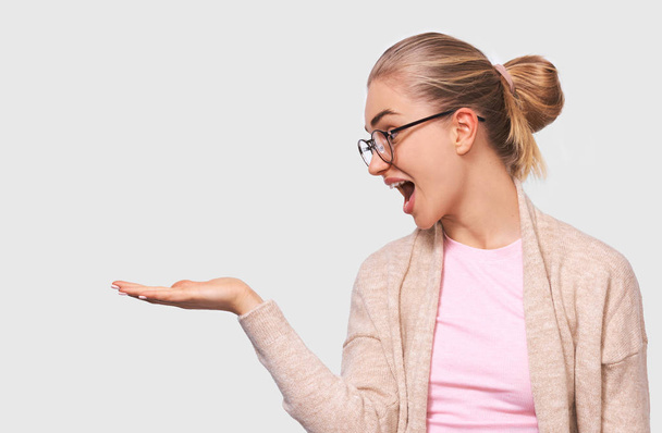 Shocked happy blonde pretty young woman looks pleasant to her palm hand, showing something in her hand, wearing round transparent eyewear and casual beige and pink blouses with blank copy space - Photo, Image