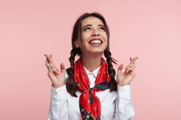 Счастливая юная леди имеет смешное выражение радости, смотрит вверх и держит пальцы скрещенными на удачу, одетый в белую рубашку и шарф
 - Фото, изображение