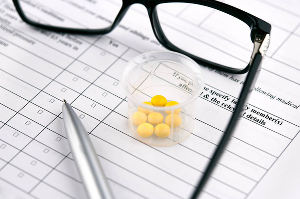 Óculos, comprimidos amarelos em vidro graduado e caneta em ques médicos
 - Foto, Imagem