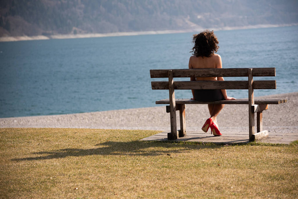 Une fille assise au-dessus de son épaule sur un banc en bois regarde un lac de montagne. La fille avec son dos à lui semble nue, portant une jupe noire et des chaussures rouges avec des talons hauts. Concept de sensualité féminine
. - Photo, image