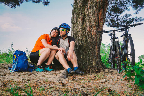 Szczęśliwa para z rowerem górskim siedzieć obejmując charakter rekreacyjny na zewnątrz. szczęśliwa para, prowadzić aktywny tryb życia, zatrzymał się w lesie w pobliżu drzewa zrobić sobie przerwę. Sport tematyczny i aktywny styl życia i romans - Zdjęcie, obraz