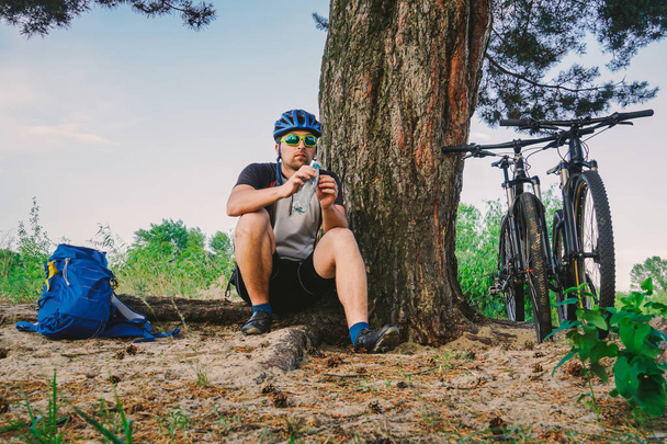 Кавказский велосипедист активный образ жизни отдыхает под деревом после занятий велосипедной питьевой водой из бутылки. Спортсмен в шлеме и спортивной одежде. Фляжка для рук. Жажда природы. Катание на горном велосипеде
 - Фото, изображение