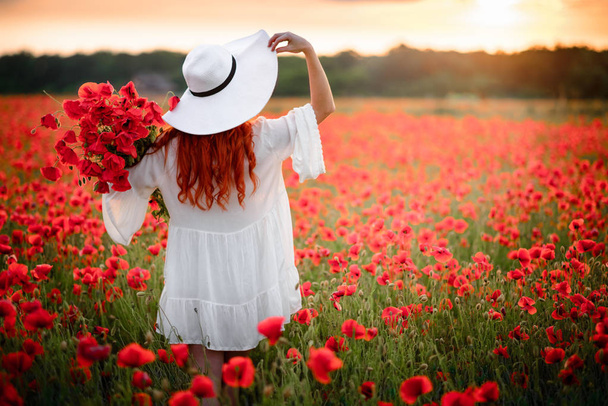 Mujer pelirroja con sombrero blanco se encuentra en el campo de flores de amapolas rojas con su espalda a la cámara
 - Foto, Imagen