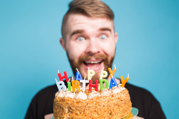 Crazy funny positive guy hipster holding a happy birthday cake in his hands standing on a blue background. Concept of jokes and holiday greetings. - Фото, зображення