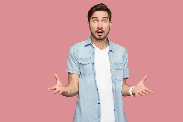 shocked sad bearded young man in blue casual style shirt standing with stressed face and looking at camera while asking on pink background - Foto, imagen
