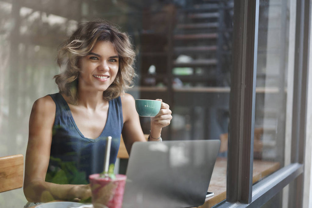 Successful lucky attractive european blond woman short curly hairstyle hold coffee cup working laptop edit photograph freelance, sit window bar cafe look outside search inspiration - 写真・画像