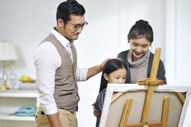 μικρό ασιατικό κορίτσι φτιάχνοντας έναν πίνακα με τους γονείς βλέποντας και αυτός - Φωτογραφία, εικόνα