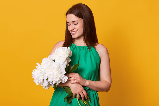 Close up portrait of attractive stylish woman on date in elegant green sundress on yellow studio background, holding peony flower bouquet, has pleasant facial expression, fells happy. Love concept. - Фото, изображение