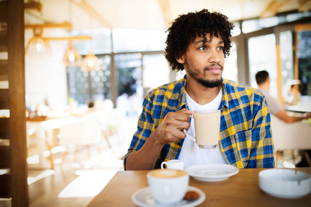 Starting new day in cafe. Side view of young african man holding coffee cup and looking away while sitting in cafe - Φωτογραφία, εικόνα