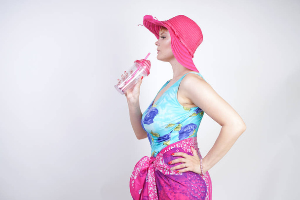 sexy plus size girl in a blue swimsuit, fuchsia hat with brim and in a trendy bright pareo stands on a white background in the Studio. a curvaceous young woman in colorful beach clothes posing alone. - Photo, Image