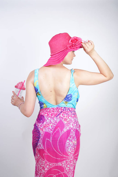 sexy plus size girl in a blue swimsuit, fuchsia hat with brim and in a trendy bright pareo stands on a white background in the Studio. a curvaceous young woman in colorful beach clothes posing alone. - Photo, Image