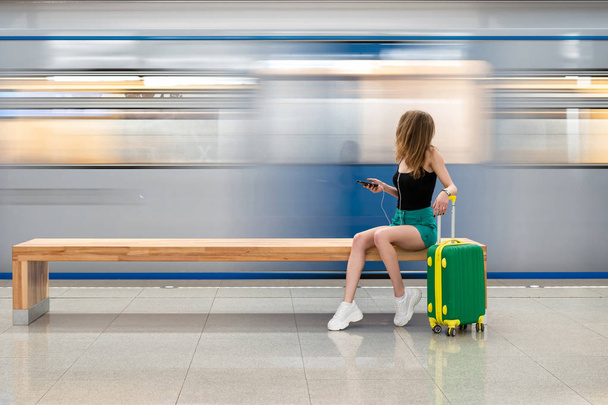 a young beautiful european girl with a small green suitcase is sitting on a bench at the railway station waiting for the train. Theme travel alone - Foto, Imagem