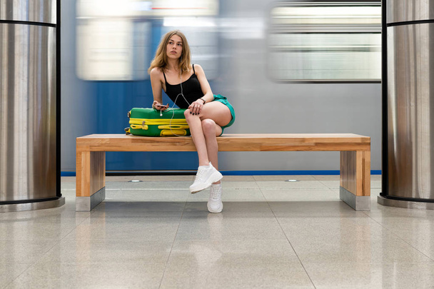 girl 25 years old sat down to relax on the bench at the subway station, train station. She travels with a green suitcase. - Photo, image