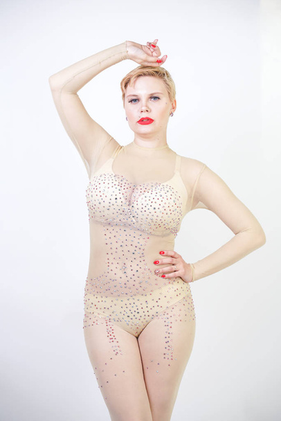 sexy curvy girl in beige lingerie and transparent overalls for whole body in rhinestones posing on white background in the Studio. fashionable young woman with short hair in erotic dance costume. - Photo, Image
