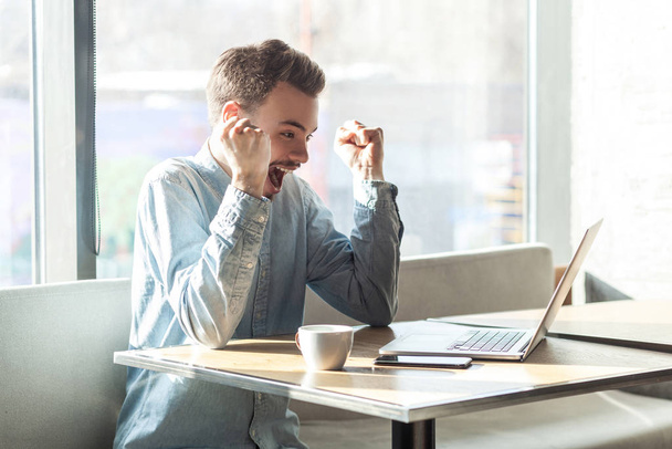 bonito espantado satisfeito positivo barbudo jovem freelancer em camisa azul olhando para tela do laptop e triunfando enquanto sentado no café
  - Foto, Imagem