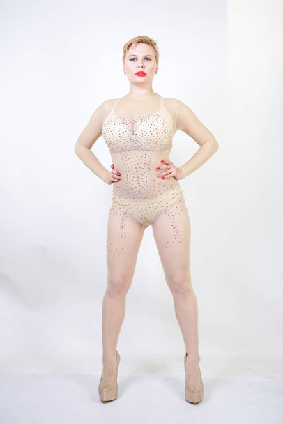 sexy curvy girl in beige lingerie and transparent overalls for whole body in rhinestones posing on white background in the Studio. fashionable young woman with short hair in erotic dance costume. - Photo, image