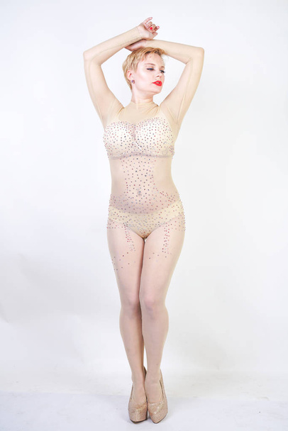 sexy bochtige meisje in beige lingerie en transparante overall voor hele lichaam in Strass poseren op witte achtergrond in de Studio. modieuze jonge vrouw met kort haar in erotische dans kostuum. - Foto, afbeelding
