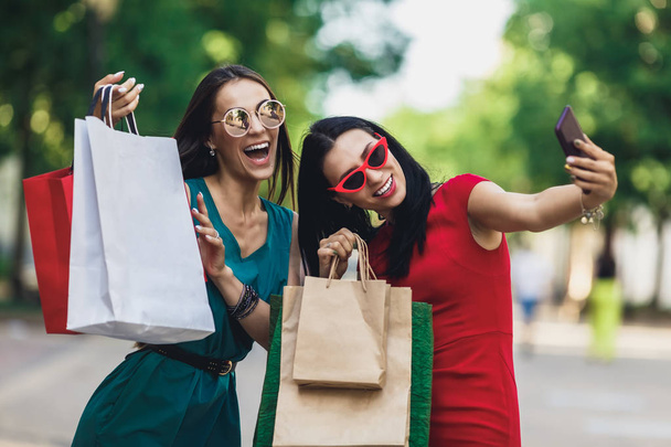 Belle ragazze felici in occhiali da sole che tengono borse della spesa, facendo selfie su smart phone e sorridendo. Concetto di vendita e consumo
. - Foto, immagini