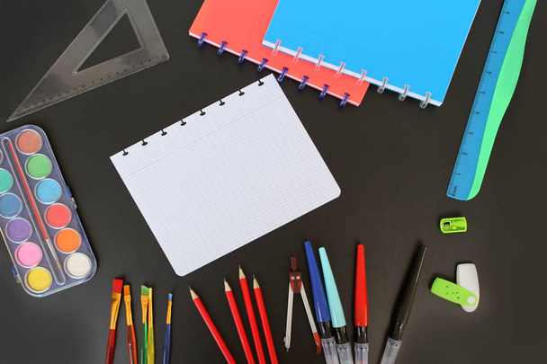 A sheet of school notebook and colored various stionionery on a black background. Back to school concept, top view, paints, pencils, pens, copy space - Photo, Image