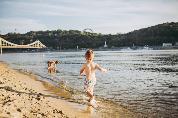 Tema verão atividades ao ar livre perto do rio na praia da cidade em Kiev Ucrânia. Pequeno menino engraçado correndo ao longo da margem do rio, saltando um respingo de água praia de areia hora de verão
 - Foto, Imagem