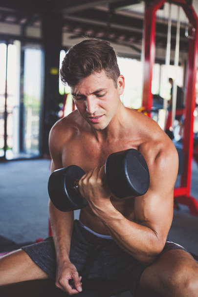handsome young man with tanned muscular naked torso sits on a bench in the gym and trains biceps with dumbbells - Foto, Bild