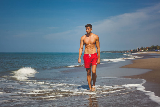 handsome sexy man in red shorts and with naked tanned fit torso walks on beach along the ocean. bright blue water and sky - Valokuva, kuva