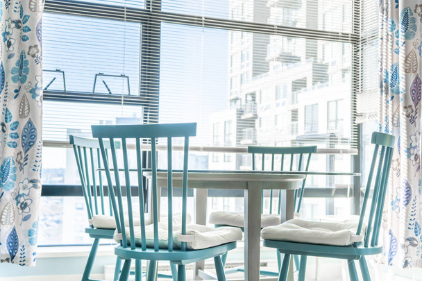 Comedor muy luminoso en un apartamento, con grandes ventanales en el fondo, y sillas azules con una mesa de cristal en primer plano. Cortinas a juego para el diseño de interiores
. - Foto, Imagen