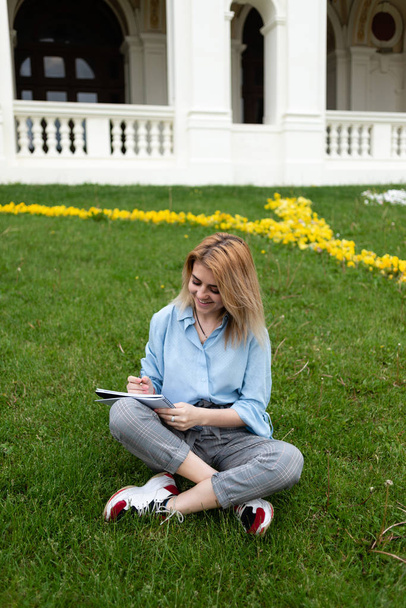 Eine glückliche Studentin sitzt auf einer Wiese in einem Park. schreibt in einem Notizbuch einen Aufsatz. - Foto, Bild